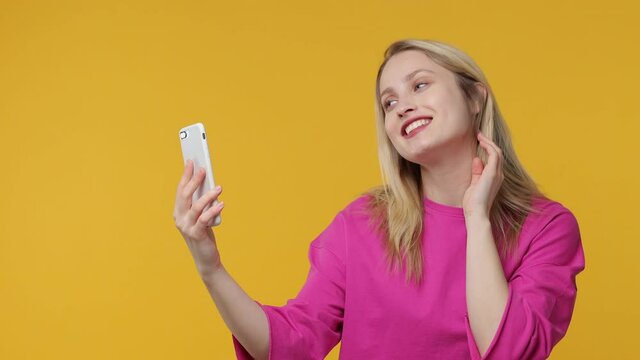 Smiling beautiful excited young woman 20s in pink t-shirt isolated on yellow background studio. People lifestyle concept. Doing selfie shot on mobile phone showing victory sign blowing send air kiss