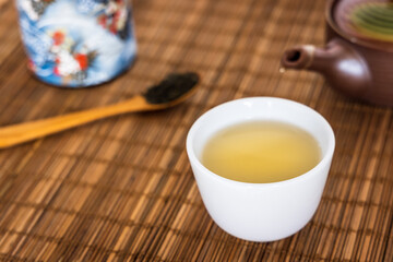 A cup of hot Japanese green tea in a white ceramic teacup put on bamboo sheet with clay porcelain teapot, tea box and full of tea leaf on wooden spoon.