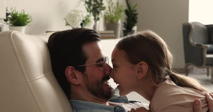 Loving 6s daughter spend weekend with caring daddy, people resting on armchair at home touch noses fooling enjoy priceless carefree time together, close up. Family bond, love, happy fatherhood concept