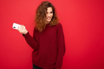 Naklejka premium Photo of pretty smiling happy young woman with curly hair wearing dark red sweater isolated on red background holding mobile phone looking to the side and having fun