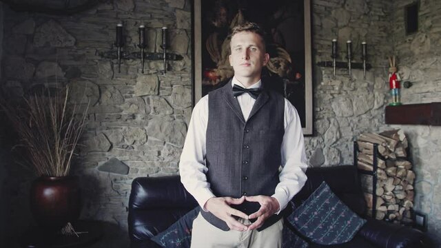 A Young Man in a Vest and Bow Tie on the Stone Wall Background Old Mansion Decorated with Paintings and Forged Candlesticks. Concept of the Wealthy Citizen of the Past Century.