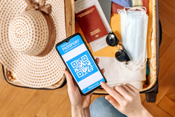 New normal concept. Close up view of female's hold a smartphone with an immune digital health passport. In the background, a suitcase with clothes and protective masks and straw hat in a blur.Flat lay