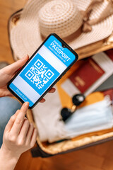 New normal concept.A female's hands hold a smartphone with an immune digital health passport. In the background, a suitcase with clothes,protective mask and straw hat in a blur. Flat lay. Vertical