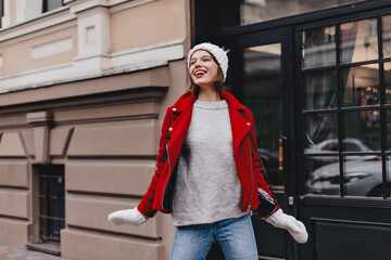 Nice girl in jeans, gray sweater, red coat and knitted hat with mittens posing with smile on street