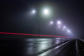 Horizontal conceptual photo of a highway illuinated by street lamps along the road at foggy nigh and red car tail light trail