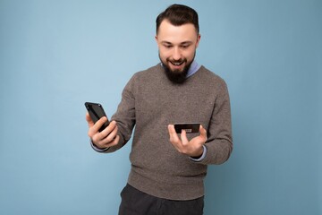 Handsome man wearing everyday clothes isolated on background wall holding and using phone and credit card making payment looking at smartphone screen