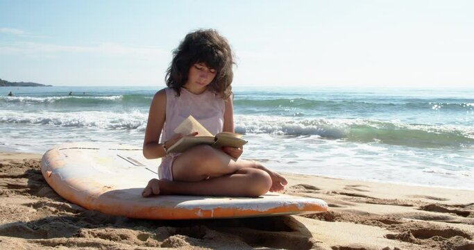 A cute girl is sitting on a surfboard on the beach and reading a book during summer vacation. 4k slow motion