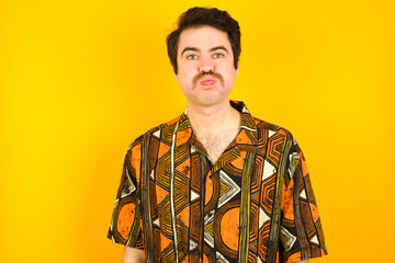 young Caucasian man wearing printed shirt against yellow wall puffing cheeks with funny face. Mouth inflated with air, crazy expression.