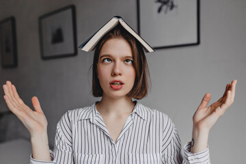 Portrait of funny girl who crosses her eyes and poses with book on her head against background of...