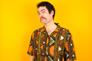young Caucasian man wearing printed shirt against yellow wall with snobbish expression curving lips and raising eyebrows, looking with doubtful and skeptical expression, suspect and doubt.