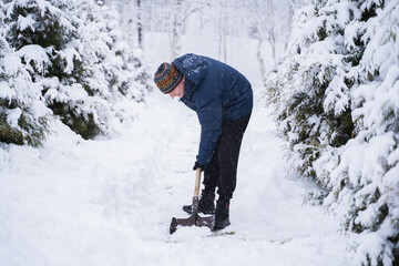 Fototapeta na wymiar Young man cleans snow from the road near the house with a large shovel. Winter, life in a country house. Warm knitted hat, jacket, boots. Alley of firs and thujas
