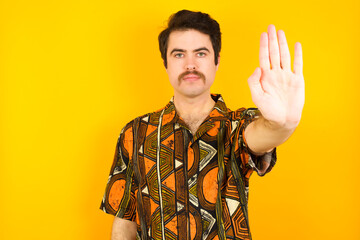 young Caucasian man wearing printed shirt against yellow wall doing stop gesture with palm of the...