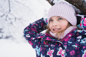 Little smiling girl, kid walking in beautiful winter forest, park among trees, branches covered with snow. Pink hat, scarf, snood, bright colorful warm jacket. Blue eyes, blond hair.Fashionable image