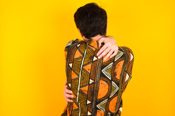 young Caucasian man wearing printed shirt against yellow wall hugging oneself happy and positive from backwards. Self love and self care.
