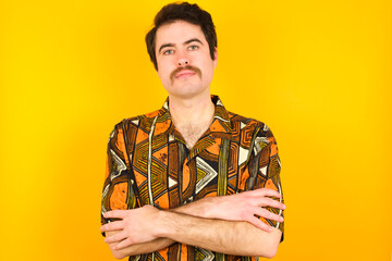 Picture of angry young Caucasian man wearing printed shirt against yellow wall looking camera.