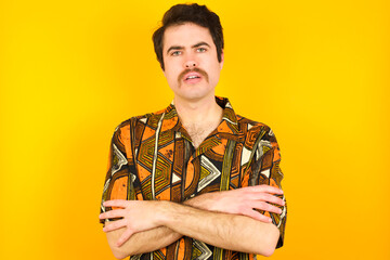 young Caucasian man wearing printed shirt against yellow wall frowning his face in displeasure, keeping arms folded, waiting for an explanation.