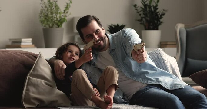 Loving father play toy guns with little preschool adorable son, people aiming shooting having fun at home, family sit on couch in cozy modern living room enjoy playtime together. Boyish hobby concept