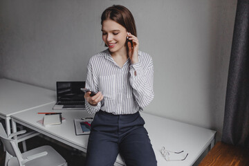 Charming woman in black pants and light blouse is sitting on table. Office worker listens to music from phone on background of laptop and stationery
