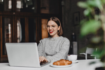 Stylish lady in glasses and cashmere sweater with smile working in gray laptop, sitting in cafe with croissant and cup of coffee on table