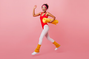 Fototapeta na wymiar Appealing african woman in aerobics outfit running on pink background. Studio shot of black fit girl with yoga mat.