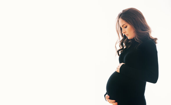 Studio Maternity Portrait Of Gorgeous Young Woman, Posing On White Background, Wearing Black Turtle Neck Dress