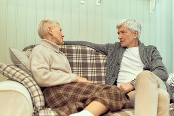 Relaxed married couple having conversation indoors. Attractive elderly man in cardigan sitting on comfortable sofa, listening to his wife, enjoying leisure tome on retirement. People and relationships