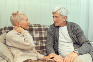 Indoor image of serious senior male with gray hair frowning, sitting on sofa with his unhappy wife, having health problems, suffering from pain in neck. People, age, retirement and health concept