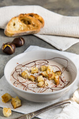 Chestnut soup with parmesan, croutons and chilli flakes and threads on grey concrete background, decorated with roasted bread and chestnuts, vertical