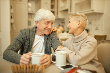 Fototapeta na wymiar Sweet portrait of charming elderly couple blonde woman and gray haired man bonding in kitchen during breakfast, touching one another, expressing love, care and affection, drinking morning coffee
