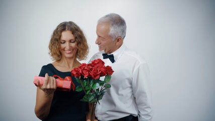 A loving elderly husband presents a gift and roses to a mature smiling woman.