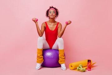 Cute girl in vintage sportswear doing exercises with dumbbells. Studio shot of curly fitness woman posing on pink background.