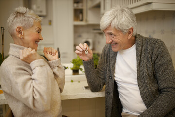 Attractive male pensioner holding keys from new house, showing them to his wife who is cheering, having overjoyed facial expression, clenching fists, excited with unexpected news. Family and home