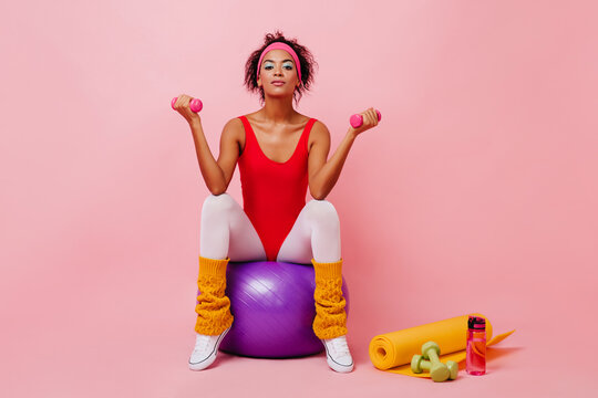 Cute girl in vintage sportswear doing exercises with dumbbells. Studio shot of curly fitness woman posing on pink background.