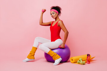 Studio shot of strong girl posing with sport equipment. Shapely african lady sitting on fitness ball.
