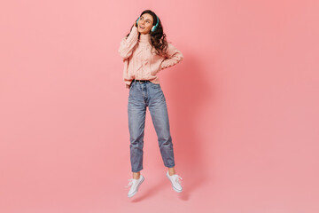 Attractive curly woman in blue moms jeans and sweater jumping on pink background, listening to song in headphones