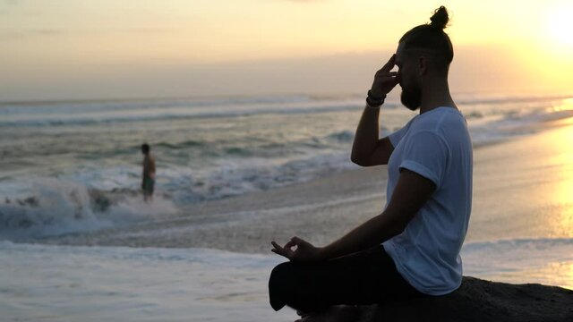 Caucasian male in lotus pose thinking about mantras recreating at coastline during perfect sunset for meditating, man training wellness vitality and inner peace of aura chakra
