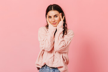 Dark-haired woman with blue eyes gently touches her face. Girl in oversized sweater and jeans looking at camera on pink background