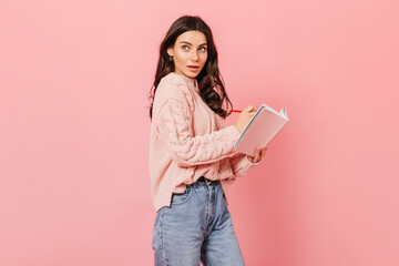 Stylish curly brunette woman posing with notebook and red pen on pink background