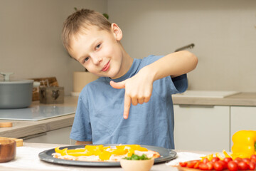 Boy 7-10 in T-shirt cooking pizza in kitchen, kid smiles and points finger at pizza