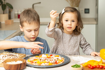 Cute little girl 2-4 in gray dress and boy 7-10 in T-shirt cooking pizza together in kitchen. Brother and sister cooking