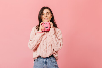 Smiling dark-haired lady in stylish sweater poses with pink camera on isolated background