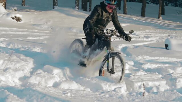 Cyclist in Black Clothes, Helmet and Snow Glasses Riding on Electric Bike in the Snow in Winter Forest. E-Bike Riding and Drifting at Cold Sunny Day - Slow Motion