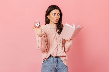 Woman is reading shocking book and holding pink alarm clock. Shot of lady in sweater and jeans on isolated background