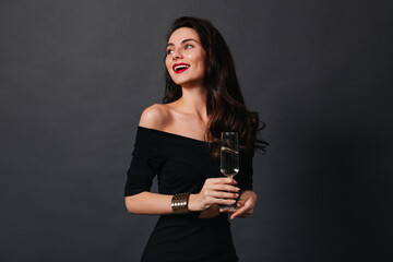 Slender dark-haired lady in small black dress and stylish gold bracelet smiles while holding glass of wine on isolated background