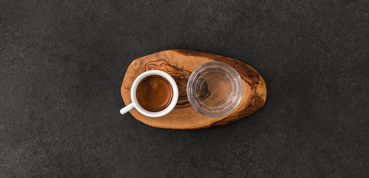 Cup of espresso coffee and glass on wooden pad on dark table, speciality coffee serving concept. Top view.