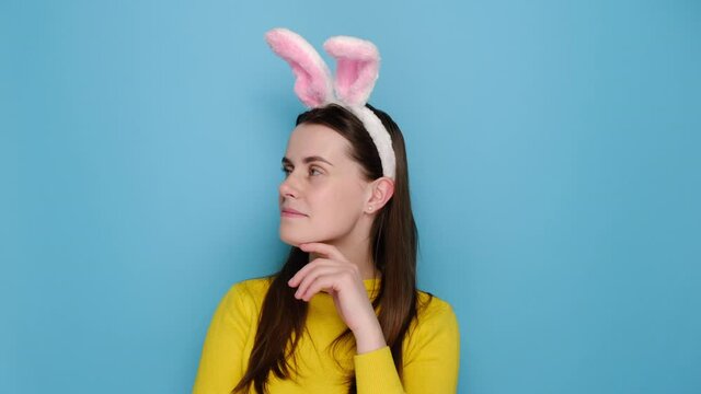 Charming young woman in pink bunny fluffy ears, is doing secret gesture, looking to camera, wears yellow sweater, standing isolated over blue studio background. People emotion and easter concept