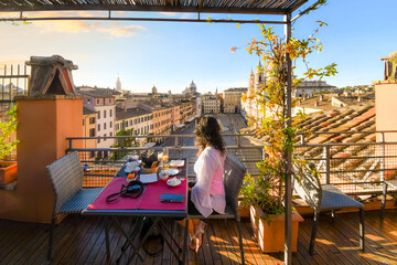A young brunette woman sits at a rooftop cafe or restaurant table enjoying the morning view of the Piazza Navona in Rome, Italy