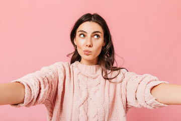 Girl in oversize sweater looks up thoughtfully. Curly brunette takes selfie on pink background and wants hugs