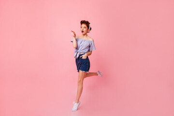 Surprised woman in denim shorts pointing with finger. Amazed pinup lady jumping on pink background.