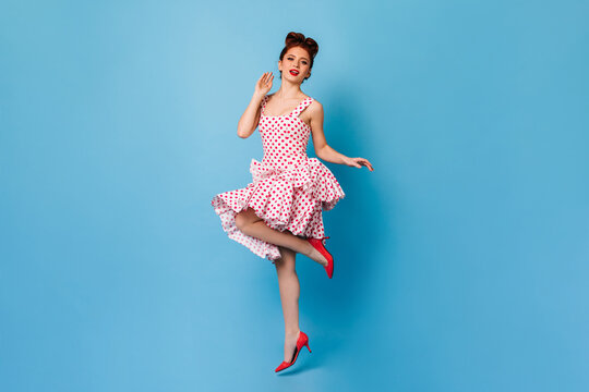 Inspired pinup girl with ginger hair standing on one leg. Studio shot of elegant woman in polka-dot dress dancing on blue background.